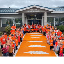 Elementary school students line the orange crosswalk in front of their school wearing orange shirts